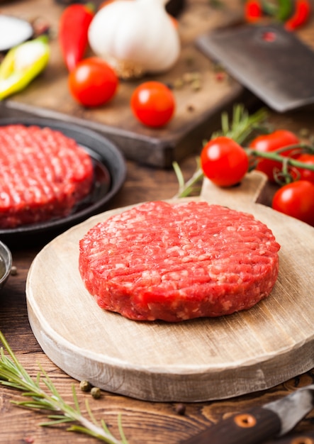 Les fermiers faits maison hachés crus frais grillent des hamburgers de boeuf sur une planche à découper ronde et une poêle avec des épices et des herbes et une hache de viande sur une planche de bois.