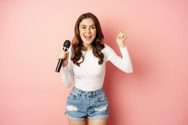 Fêtarde. Heureuse jeune femme chantant dans le microphone, interprétant une chanson, s'amusant lors d'un événement, debout sur fond rose.