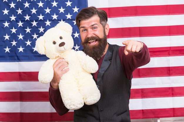 Fête américaine. Hipster barbu homme tenir fond de drapeau américain ours en peluche. Garde d'enfants. Encore enfantin. Nourrisson et drôle. 4 juillet. Le jour de l'indépendance. Un type américain avec un jouet célèbre les vacances.