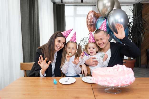 Fête d'anniversaire et famille en chapeaux de vacances à la table en agitant la caméra. Gâteau rose et ballons.