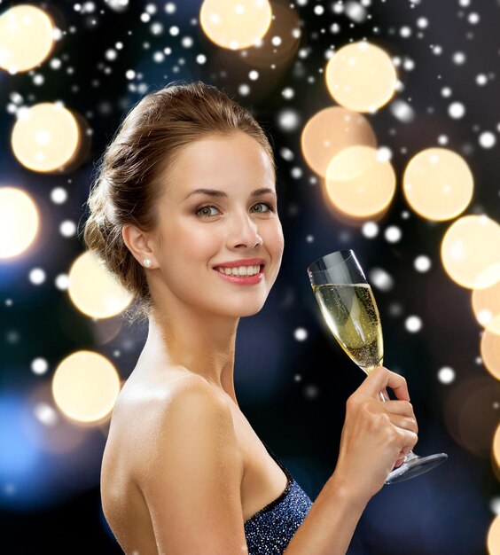 fête, boissons, vacances, noël et concept de personnes - femme souriante en robe de soirée avec un verre de vin mousseux sur les lumières de la nuit et sur fond de neige