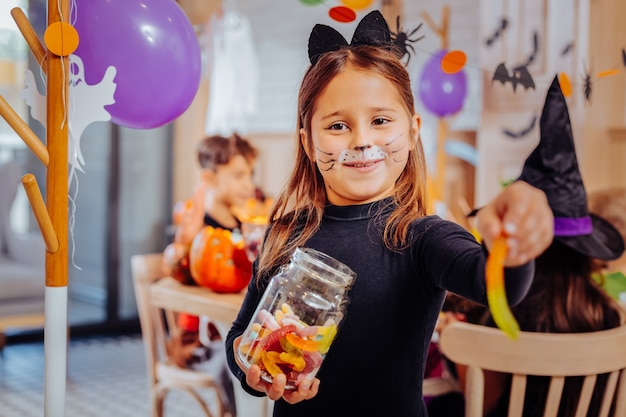 Fête d'Halloween. Belle jolie fille aux yeux sombres portant un costume de chat mangeant un ver gommeux tout en assistant à la fête d'Halloween