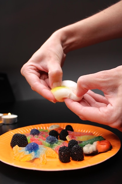 Photo fête d'halloween les mains brisent un œil de gelée blanc et jaune plaque avec des bonbons et des bonbions bougie