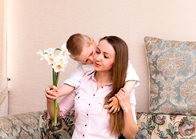 Fête des mères, vacances et concept de famille - un petit fils heureux donne des fleurs à sa mère souriante à la maison. Un enfant offre à maman un bouquet de jonquilles