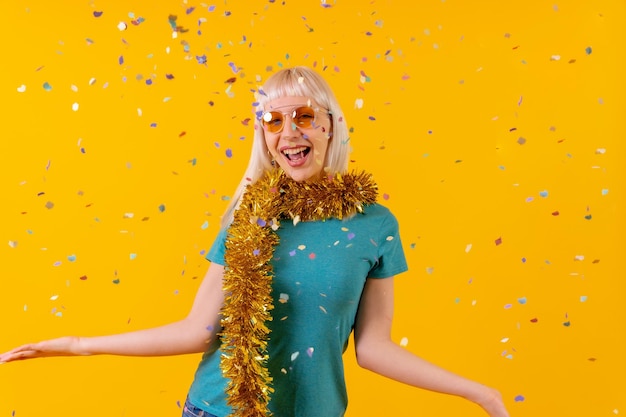 Fête souriante avec des confettis lors d'une célébration fille caucasienne blonde en studio sur fond jaune