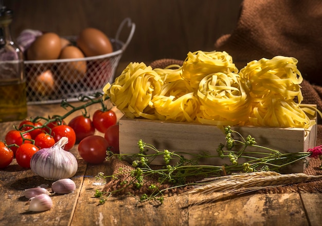 Fettuccine de pâtes sur table en bois avec tomates cerises, poivre, farine de blé et œufs en arrière-plan.