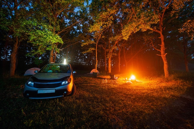 Feu de camp nocturne dans la forêt près d'une voiture argentée et de deux tentes et chaises portables