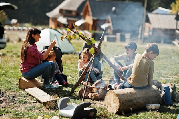 Feu de joie familial en montagne. Faire frire des saucisses. Mère avec quatre enfants en camping. Randonnée d'automne et météo du camp. Réchauffement et cuisson près de la flamme ensemble.