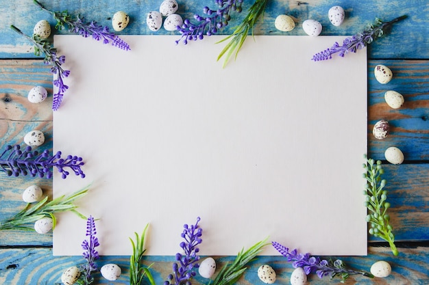 Une feuille de papier blanc encadrée de fleurs violettes et d'œufs de caille sur une table en bois bleu usé