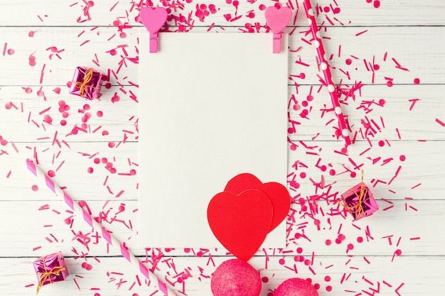 Feuille de papier blanc vide avec des décorations de confettis et de valentines sur une vue de dessus en bois blanc. Composition de vacances