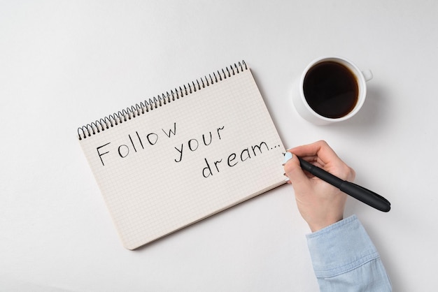 Feuille de papier avec les mots Suivez votre rêve Femme main écrit dans une tasse de café pour ordinateur portable Vue de dessus fond blanc