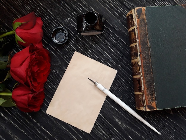 Feuille de papier vierge avec un stylo plume, un pot d'encre, un livre ancien et trois roses rouges