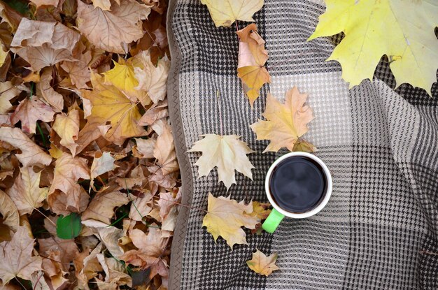 Photo les feuilles d'automne et la tasse de café chaude à la vapeur se trouvent sur un plaid à carreaux à l'extérieur.