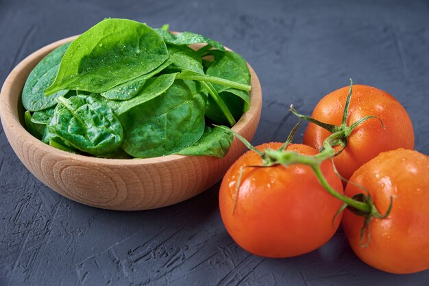 Feuilles d'épinards frais dans un bol en bois et tomate sur fond sombre.