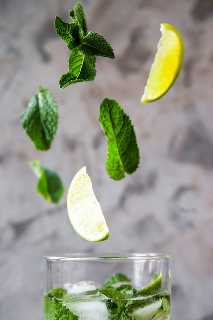 Les feuilles de menthe et de citron vert volent dans un verre avec du mojito avec de la glace sur un fond gris