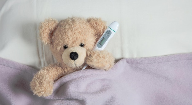 Fever cold flue Cute teddy in bed recouvert d'une couverture chaude tenant un thermomètre