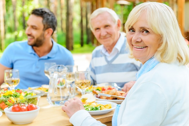 Fier d'avoir une grande famille. Famille heureuse assise à la table à manger à l'extérieur tandis qu'une femme âgée regarde par-dessus l'épaule et sourit