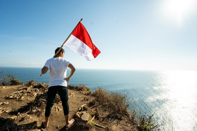 Fier homme indonésien sur une falaise de plage soulevant le drapeau de l'Indonésie rouge et blanc