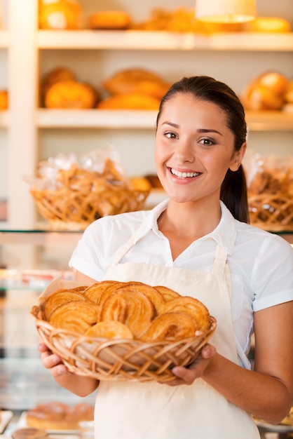 Fier de ses pâtisseries. Belle jeune femme en tablier tenant un panier avec des produits de boulangerie en se tenant debout dans une boulangerie