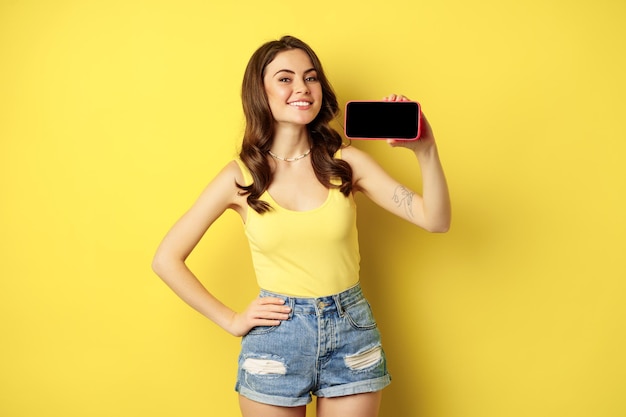 Fière femme souriante montrant l'écran horizontal du téléphone portable, l'air fier et satisfait, démontrant l'affichage du smartphone, l'application ou la boutique en ligne, fond jaune.