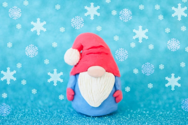 Figurine de carte de Noël d'un gnome drôle avec une barbe et un gros nez sur fond bleu et des flocons de neige qui tombent.