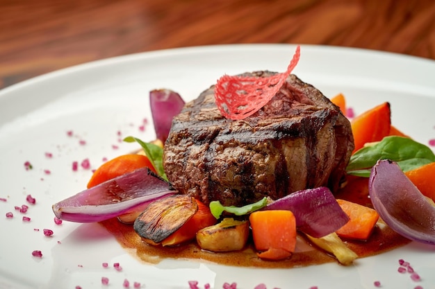 Filet mignon de boeuf aux oignons cuits au four et légumes dans une assiette blanche sur un fond en bois