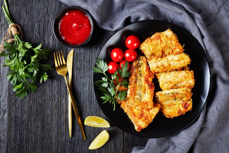 Photo filet de poisson de merlu frit croustillant servi sur plaque noire avec quartiers de citron, tomates cerises, ketchup, persil frais sur une surface en bois sombre, gros plan, plat, vue de dessus