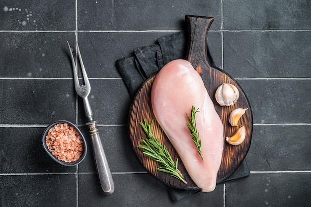 Filet de poitrine de poulet cru sur une planche de bois avec de la viande de volaille au romarin et à l'ail Fond noir Vue de dessus