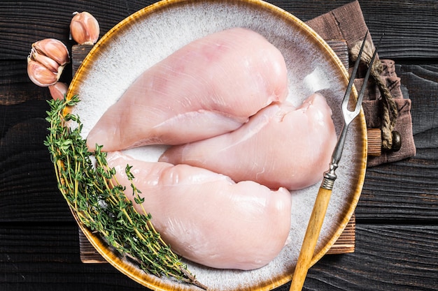 Filet de poitrine de poulet viande crue dans une assiette avec des herbes Fond en bois noir Vue de dessus