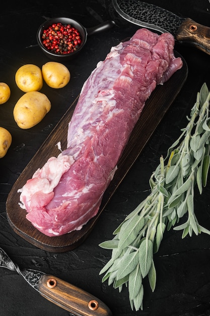 Photo filet de porc cru à la sauge, sur une planche à découper en bois, avec un couteau à barbecue et une fourchette à viande, sur une table en pierre noire, vue de dessus à plat