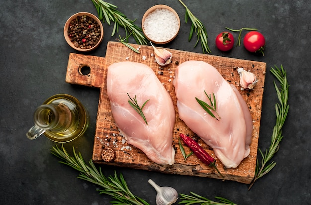 Filet de poulet cru sur une planche à découper avec du romarin, des épices, des tomates et du poivre sur un fond de pierre