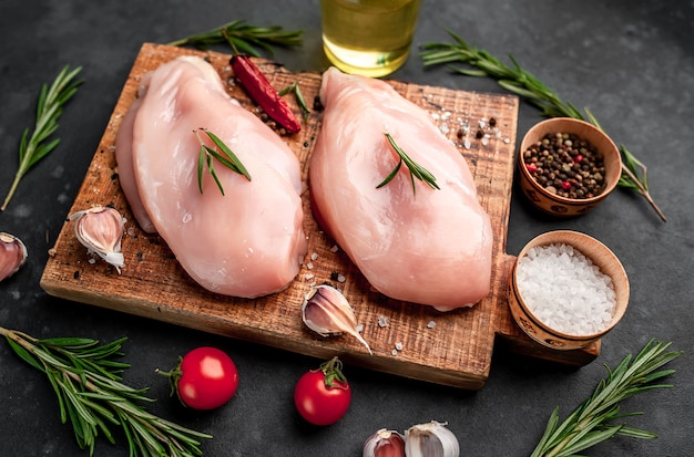 Filet de poulet cru sur une planche à découper avec du romarin, des épices, des tomates et du poivre sur un fond de pierre