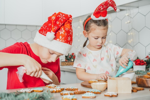 Fille de 3 ans et garçon de 8 ans en bonnet de Noel décorent des biscuits en pain d'épice avec du glaçage. Frères et sœurs dans la cuisine de Noël