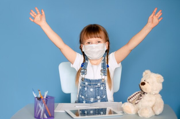 Une fille de 5 à 6 ans assise à une table effectue des tâches à distance sur la tablette. Isoler sur un mur bleu. Coronavirus, enfant masqué.