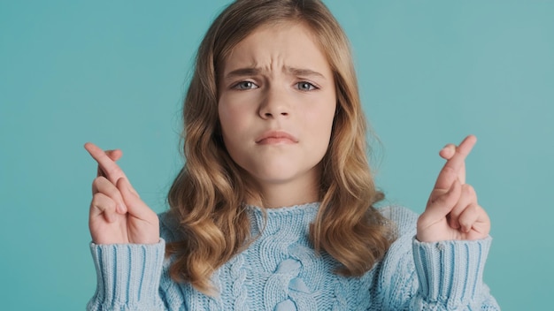 Fille adolescente blonde bouleversée à la recherche d'inquiétude en gardant les doigts croisés devant la caméra sur fond bleu. Expression de prière