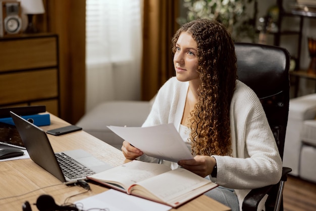 La fille adolescente dans un chandail blanc étudie en ligne depuis la maison est assise sur une chaise de bureau