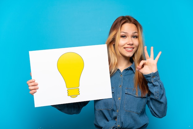 Photo fille adolescente sur fond bleu isolé tenant une pancarte avec icône ampoule avec signe ok