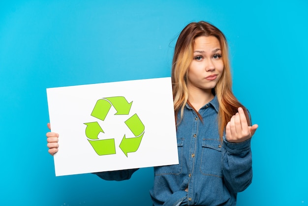 Fille adolescente sur fond bleu isolé tenant une pancarte avec l'icône de recyclage et faire le geste à venir