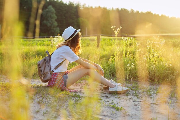 Fille adolescente heureuse au chapeau avec sac à dos assis sur une route de campagne, profitant des vacances d'été, nature pittoresque, espace de copie