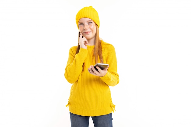 Fille adolescente heureuse aux cheveux rouges, sweat à capuche jaune et pantalon communiquent avec téléphone et sourires isolés sur fond blanc