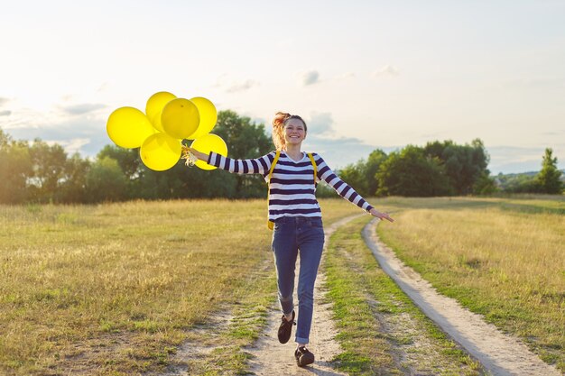 Fille adolescente heureuse avec des ballons jaunes et un sac à dos courant et sautant le long d'une route de campagne dans une prairie d'été. Liberté, vie, joie, concept de vacances