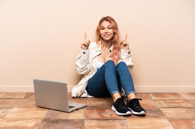 Fille adolescente avec un ordinateur portable assis sur le sol à l'intérieur pointant vers le haut une excellente idée