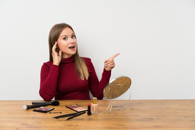 Fille adolescente avec palette de maquillage et cosmétiques dans une table surprise et pointant le doigt sur le côté