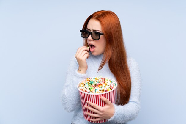 Fille adolescente rousse sur bleu avec des lunettes 3d et tenant un grand seau de pop-corn tout en regardant de côté