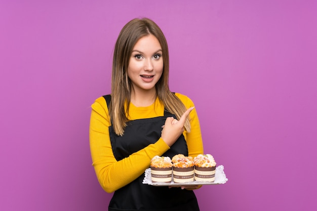 Fille adolescente tenant beaucoup de mini gâteaux différents sur un mur violet isolé pointant le doigt sur le côté