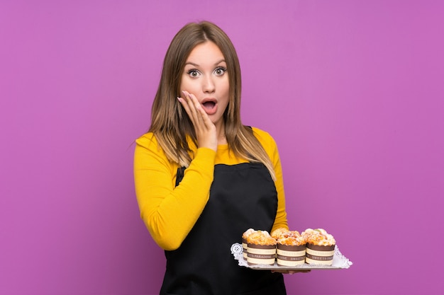 Fille adolescente tenant plein de mini gâteaux différents avec une expression du visage surprise