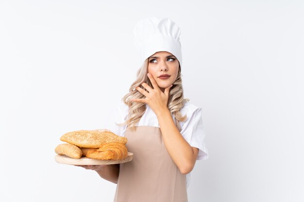 Fille adolescente en uniforme de chef. Femme boulanger tenant une table avec plusieurs pains sur fond blanc isolé, pensant à une idée