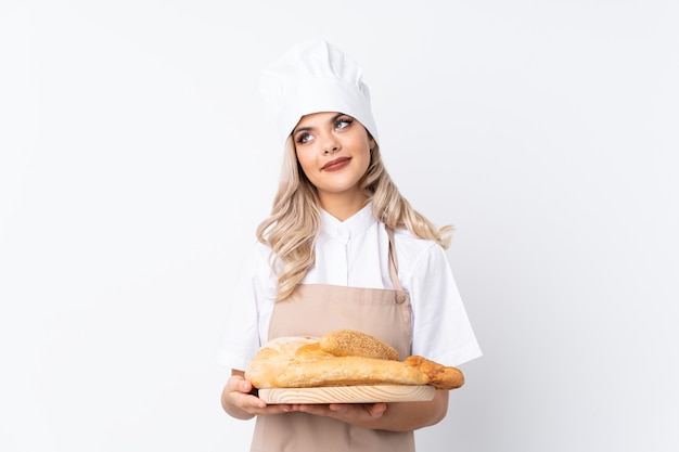 Fille adolescente en uniforme de chef. Femme boulanger tenant une table avec plusieurs pains sur fond blanc isolé en riant et levant