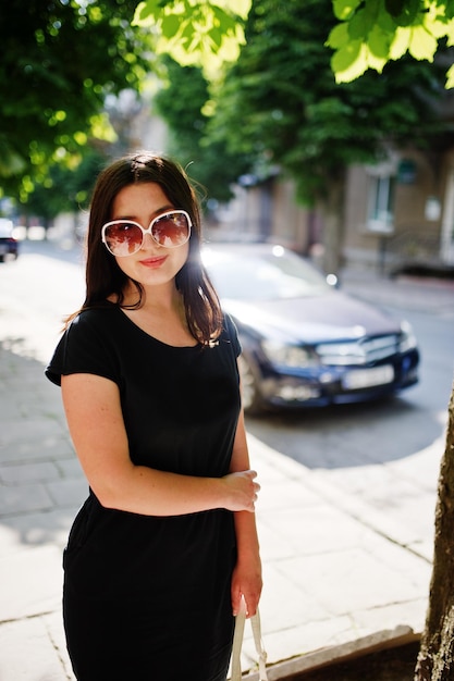 Fille d'affaires brune en robe noire sur des lunettes de soleil avec sac à main à la main posant dans la rue de la voiture de fond de la ville sur la route