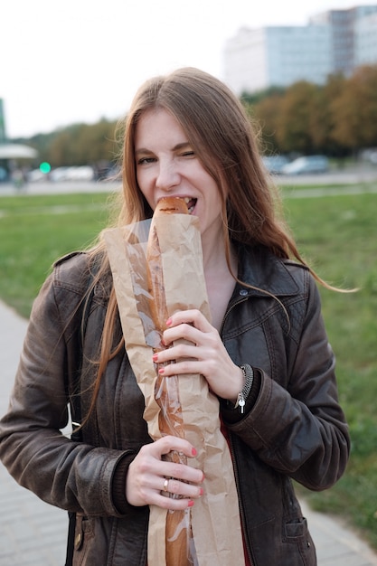 Fille affamée mordant un long pain en marchant dans la rue